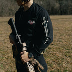 Person in tactical gear holding a rifle in an open field with trees in the background