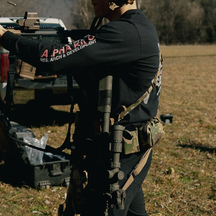 Person in tactical gear holding a rifle in an open field with a truck in the background.