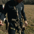Person in tactical gear holding a rifle in an open field with trees in the background