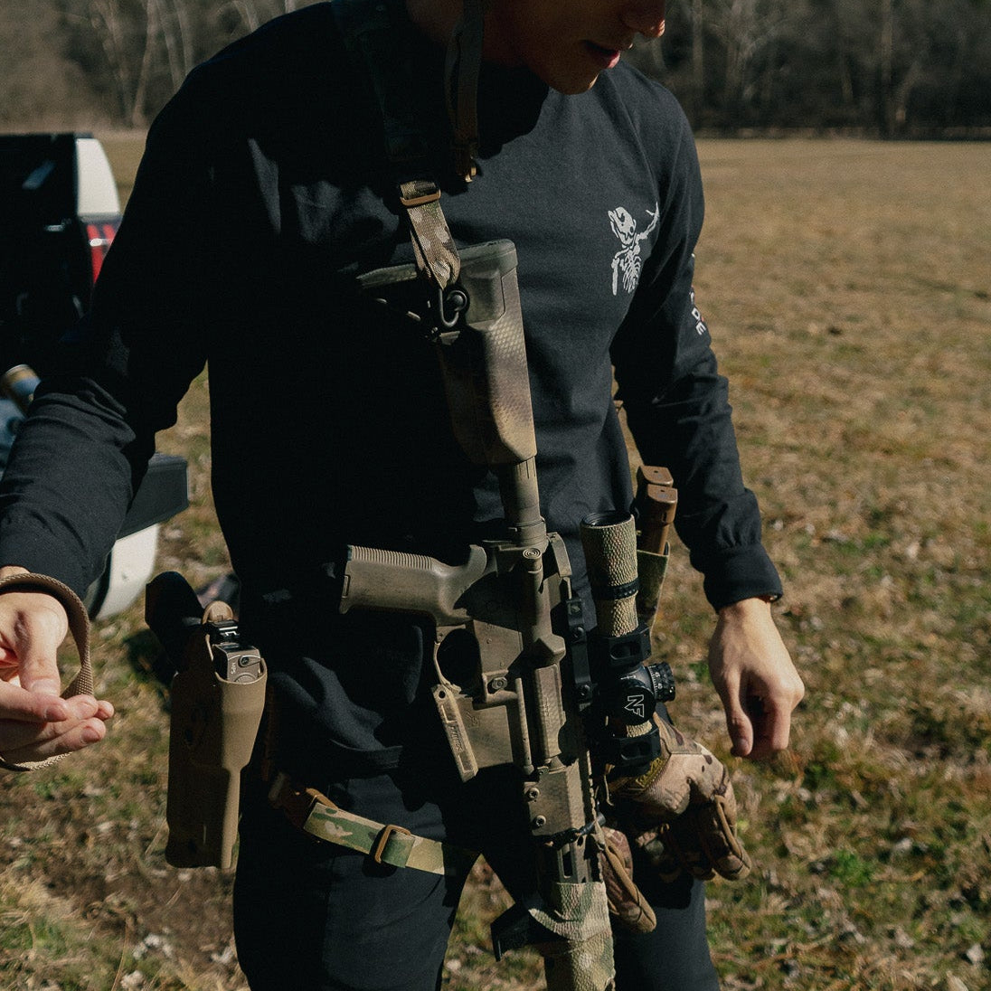 Person in tactical gear holding a rifle in an open field with trees in the background