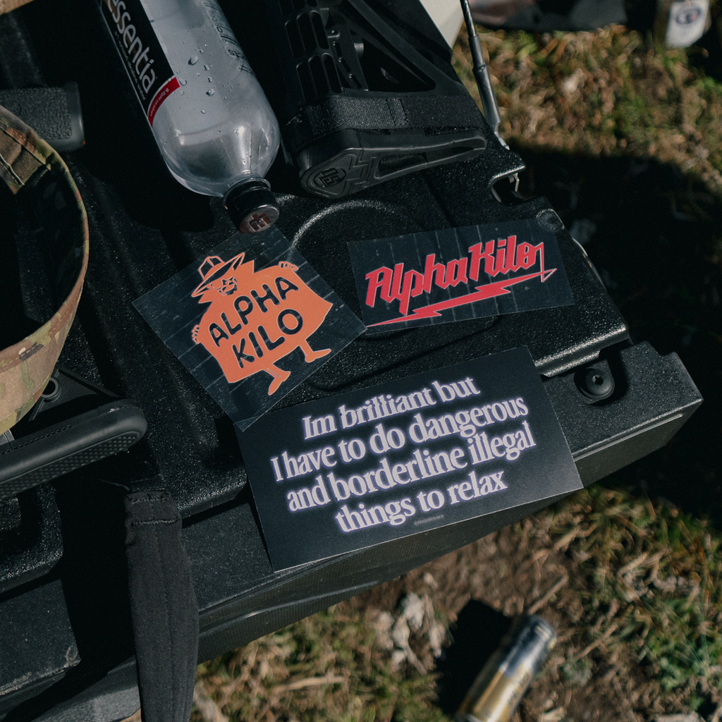 Close-up of a machine gun with 'Alpha Kilo branding and text on a grassy background.