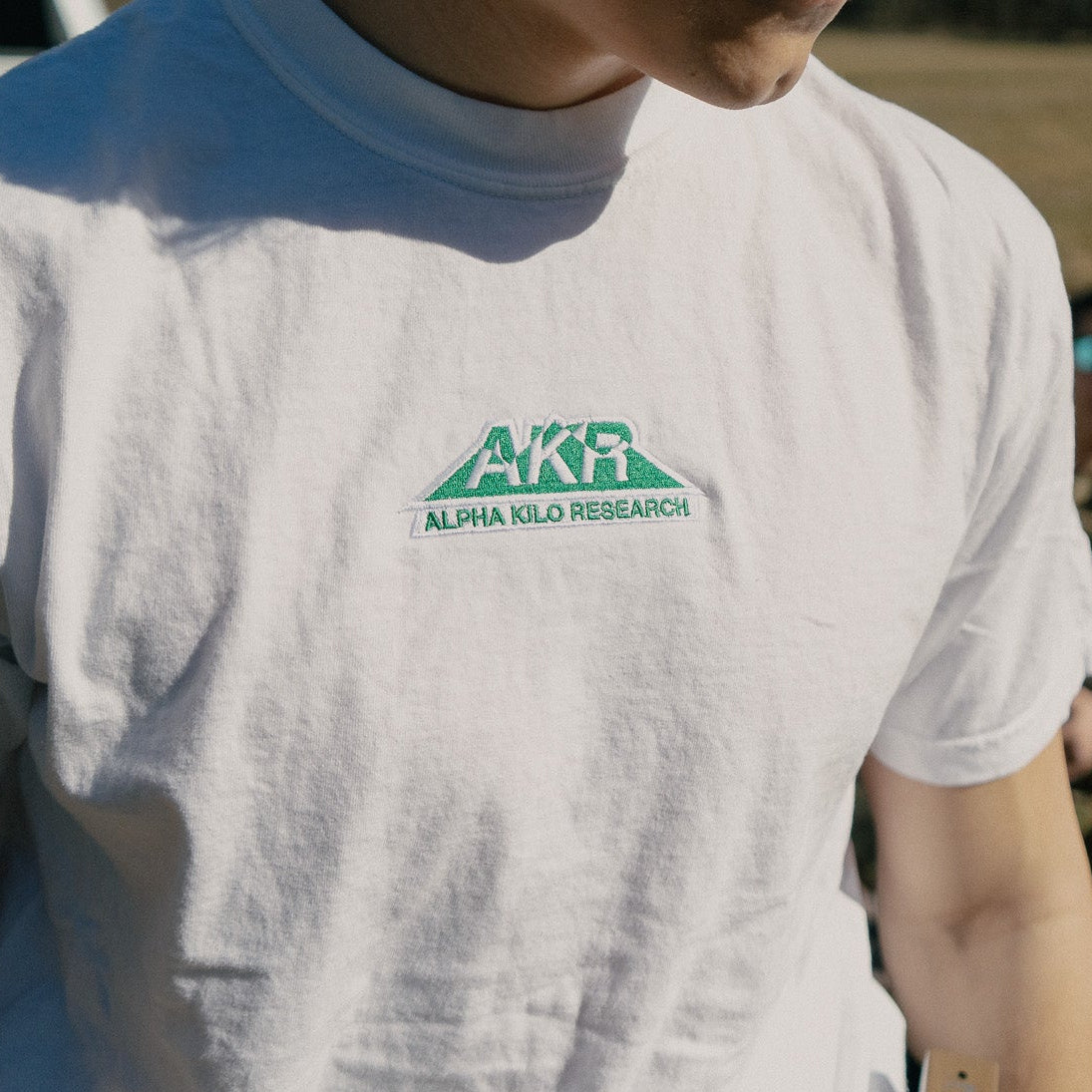 Person wearing a white t-shirt with 'AKR' logo in an outdoor setting
